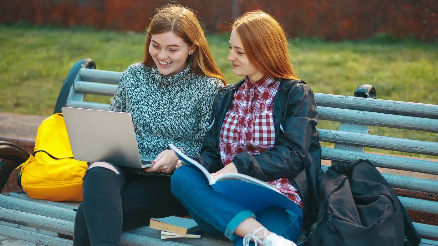 To unge piger sidder på en bænk i en park og kigger på en bærbar computer